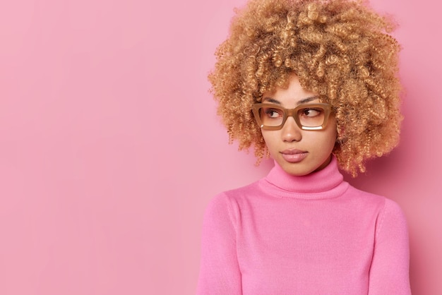 Free photo studio shot of serious curly haired young european woman looks away with concentrated expression considers something dressed in casual clothes isolated over pink background blank copy space away.