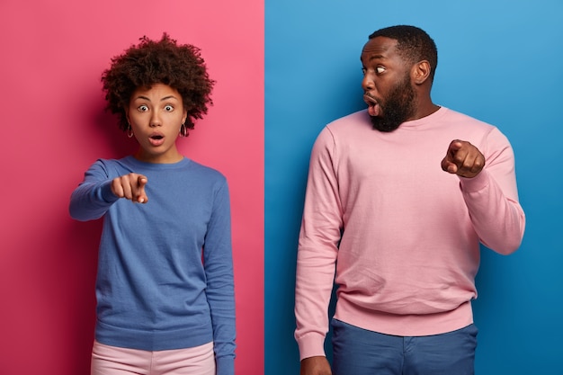 Free photo studio shot of shocked ethnic woman and man notices very strange object, points directly at camera, cannot believe eyes