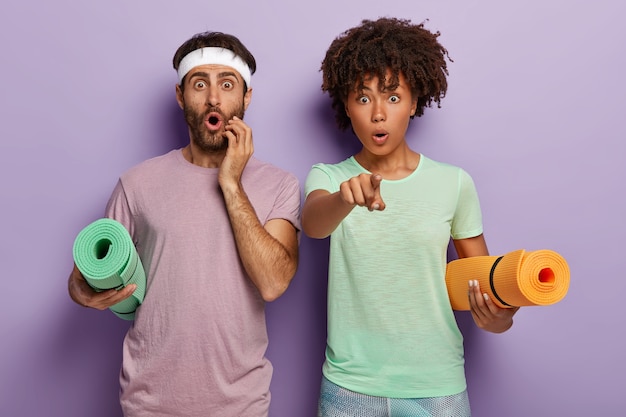 Free photo studio shot of sporty woman and man holds yoga mats, shocked and stares straightly at camera