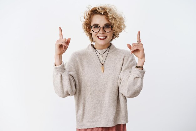 Studio shot of stylish queer girl with short blond hair lipstick and glasses pointing up with raised hands and enthusiastic smile showing cool copy space for promotion over white wall.