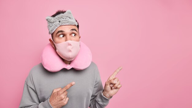 Studio shot of surprised adult man wears sleepmask protective mask on face travel pillow around neck shocked by something unexpected points away on blank space 