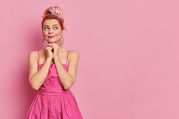 Free Photo studio shot of thoughtful dreamy expression has bright makeup keeps hands under chin prepares for carnival party wears crown and pink dress stands indoor with blank space for your promotion.