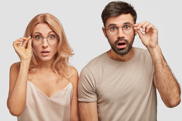 Studio shot of two surprised terrific woman and man look in bewilderment, touch rim of spectacles, being amazed by sudden news