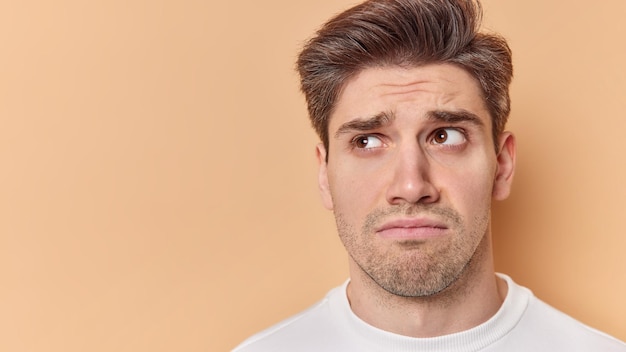 Studio shot of unhappy brunet young man has sulking expressionn looks sadly away feels disappointed has some problems isolated over brown background with blank space for promotional content.