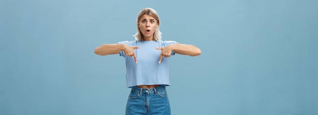 Studio shot of worried caucasian young woman feeling nervous her shoes do not fit frowning asking advice nervously pointing down shocked while frowning and standing over blue background