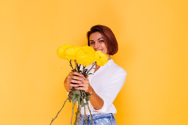 Free photo studio shot on yellow background happy caucasian woman short hair wearing casual clothes white shirt and denim pants holding bouquet of yellow asters