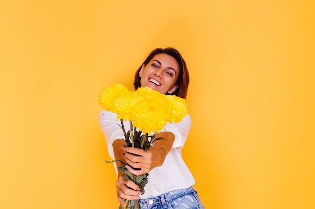 Free photo studio shot on yellow background happy caucasian woman short hair wearing casual clothes white shirt and denim pants holding bouquet of yellow asters