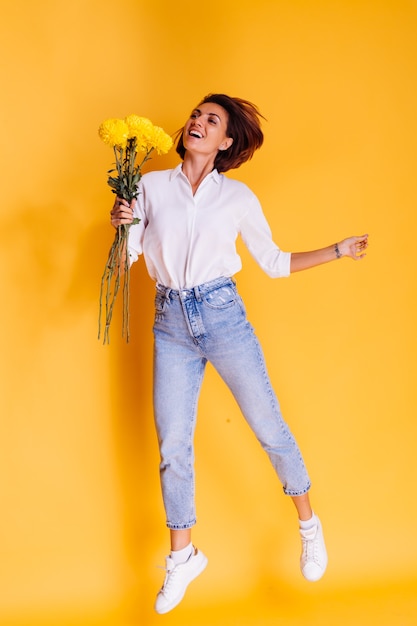 Studio shot on yellow background Happy caucasian woman short hair wearing casual clothes white shirt and denim pants holding bouquet of yellow asters