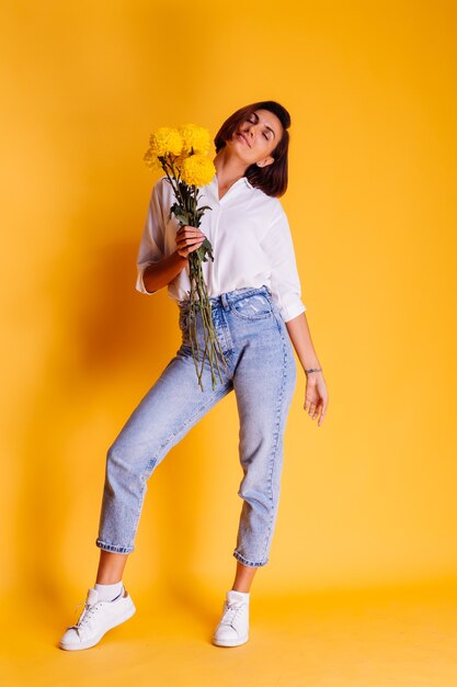 Studio shot on yellow background Happy caucasian woman short hair wearing casual clothes white shirt and denim pants holding bouquet of yellow asters