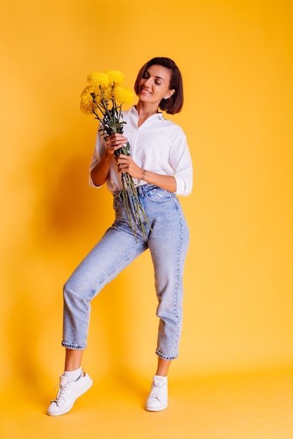 Studio shot on yellow background Happy caucasian woman short hair wearing casual clothes white shirt and denim pants holding bouquet of yellow asters