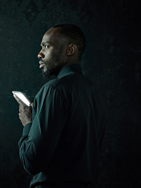 Studio shot of young serious black African man thinking while talking on mobile phone against black studio background