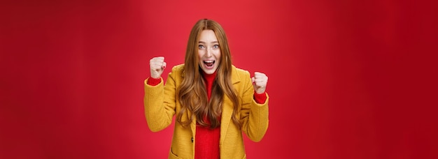 Free photo studio shto of cheerful excited goodlooking ginger girl in yellow coat raising clenched fists in joy