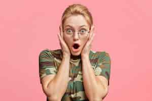 Free photo stunned blonde female model in eyewear, being astonished with something, keeps mouth opened, dressed in camouflage t shirt, isolated over pink