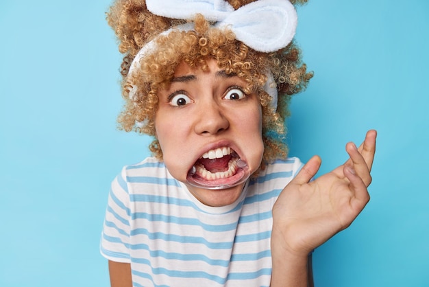 Stunned emotional curly haired woman stares bugged eyes uses mouth expander keeps palm raised wears headband and striped t shirt reacts on something embarrassing isolated over blue background