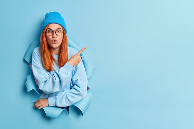 stunned redhead young woman points at upper right corner demonstrates copy space with surprised expression dressed in fashionable youth clothes.