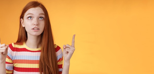 Free Photo stunned thrilled young redhead woman peer focused pointing up index fingers upwards look concentrated excited hold breath amused performance standing orange background intrigued and curious