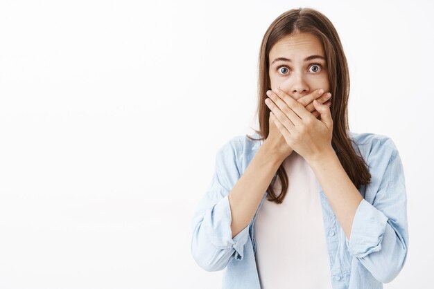 Stunned worried european female in blue shirt over t-shirt gasping covering mouth with palms from amazement popping eyes