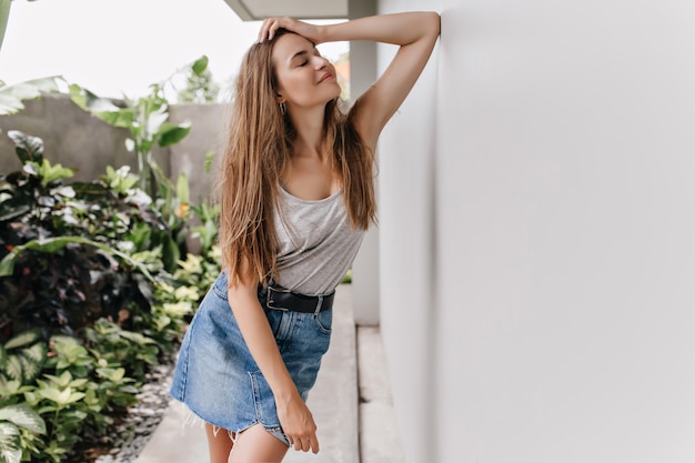 Stunning slim girl posing near plants with inspired face expression. Magnificent lady in retro denim skirt chilling.