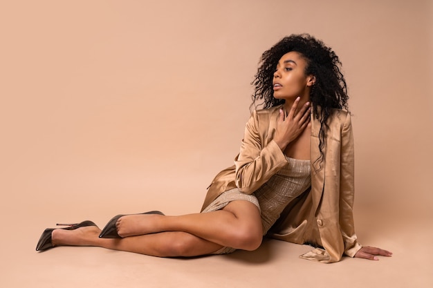 Stunning tanned model with voluminous curly hairstyle in shiny golden dress and satin blazer sitting on beige floor