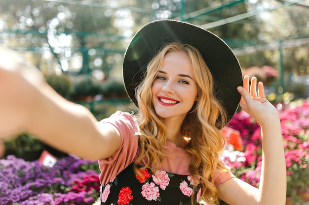 Stunning woman with beautiful eyes making selfie on orangery. Pleased woman in black hat posing with flowers.