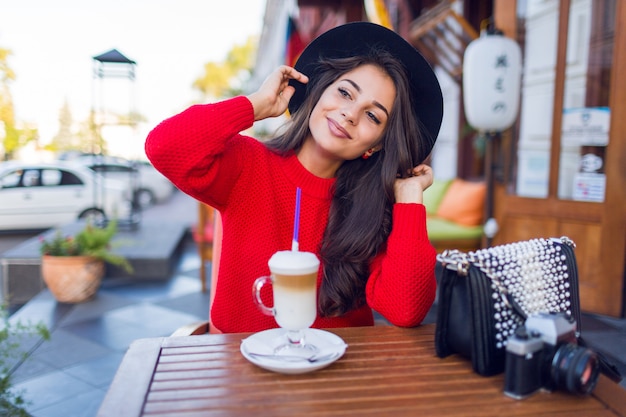 Free photo stunning young lady in stylish black hat and bright red sweater sitting in open space cafe and drink coffee with milk or cappuccino.