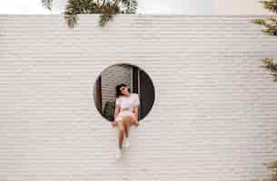 Free photo stunning young woman sitting on bricked wall outdoor shot of adorable girl in sunglasses posing on urban background