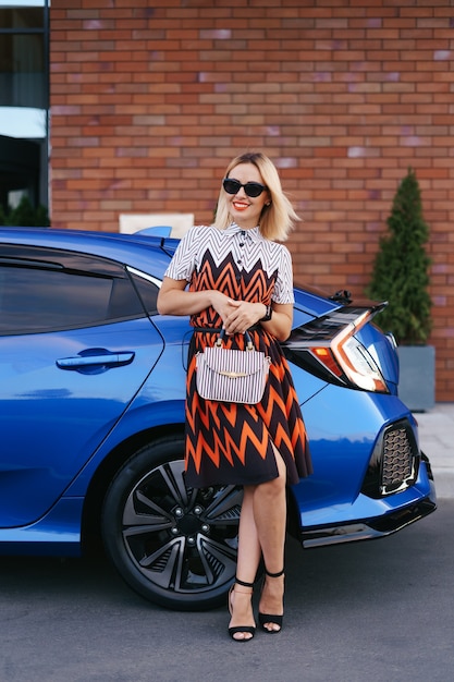 Free photo stunning young woman waring dress posing in front of her car outdoors, ownership driver