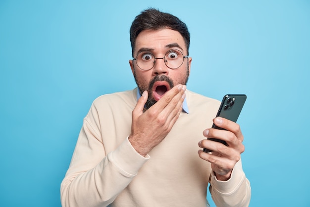 Stupefied bearded adult European man keeps mouth opened stares terrified, holds mobile phone finds out shocking news dressed in casual jumper 