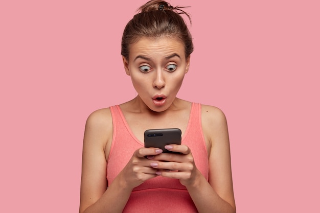 Stupefied emotional woman stares with bugged eyes at smart phone, feels shocked as reads news at web page, connected to wireless internet, has break after training with coach in gym, wears pink top