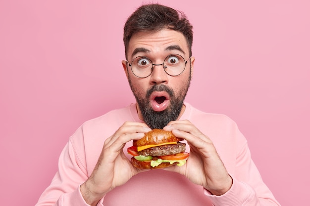 Free photo stupefied handsome man poses with delicious appetizing fast food holds tasty hamburger wears round spectacles casual jumper doesnt keep to diet likes cheat meal dressed casually