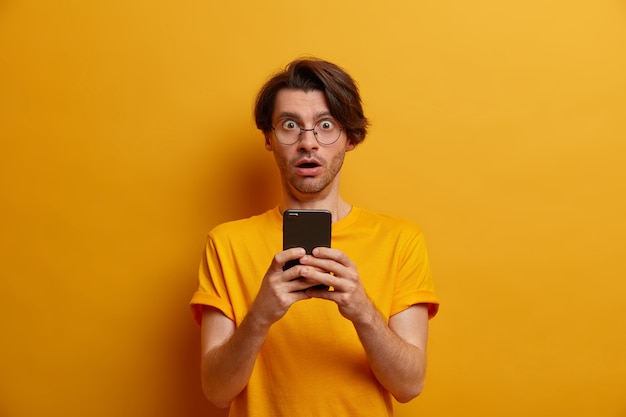 Stupefied man distracted from screen of smartphone, watches horrible video, gasps from wonder, got unexpected message, wears round sprectacles and t shirt, isolated over vivid yellow wall