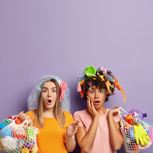 Free Photo stupefied two female activists stare with omg expression, shocked to pick up much garbage, hold net bags with plastic waste, dressed in casual outfit, pick up litter for recycling, free space above