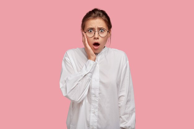 Stupefied young woman with glasses posing against the pink wall