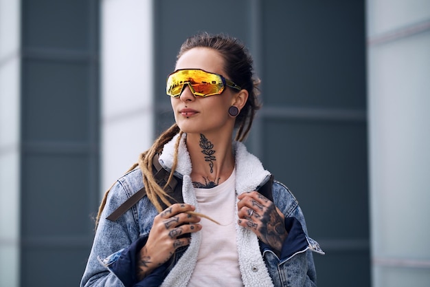 Free photo styilish punk girl wirh dreadlocks, tatooes on her hands and big toned glasses is standing over skyscraper.