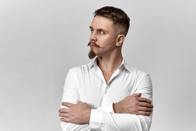 Style, fashion and business concept. Studio image of confident elegant young European businessman with stylish mustache and beard crossing arms on his chest and looking away, having thoughtful look