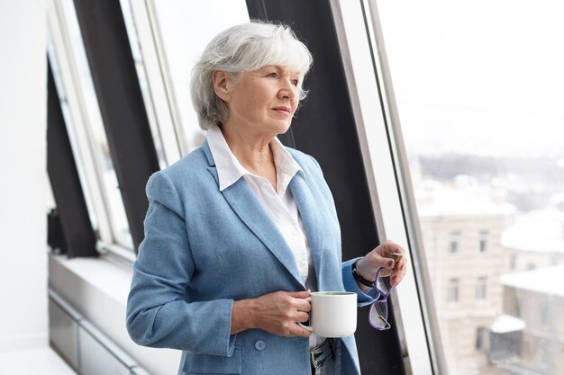 Style, fashion, career and age concept. Successful elegant gray haired female in her sixties holding glasses and mug, drinking coffee and looking through window, having pensive thoughtful expression