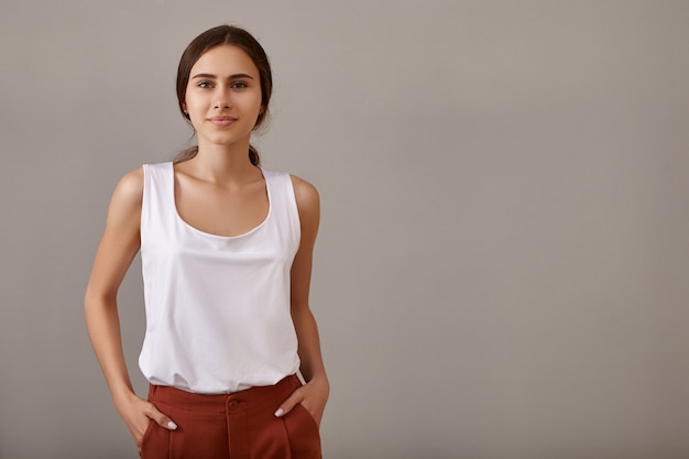 Style, fashion, elegance and confidence concept. Stylish young European woman with bare tanned shoulders posing isolated against blank copyspace wall, keeping her hands in pockets