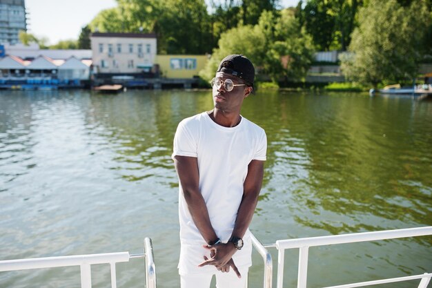 Stylish african american boy wear on white clothes glasses and cap against pier of lake Street fashion of young black people