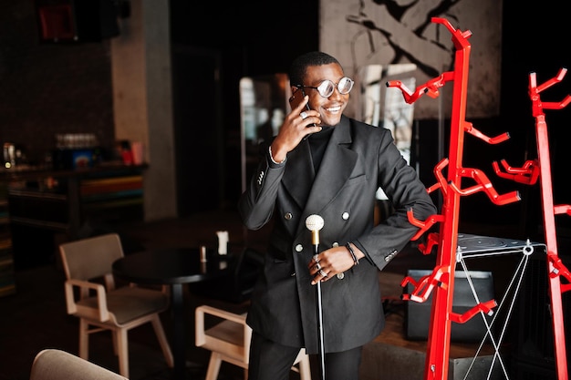 Free photo stylish african american gentleman in elegant black jacket and glasses holding retro walking stick as cane flask or tippling cane with golden diamond ball handle and speaking on mobile phone