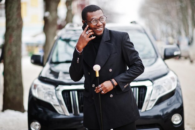 Stylish african american gentleman in elegant black jacket holding retro walking stick as cane flask or tippling cane Rich fashionable afro man against business suv car speaking on mobile phone