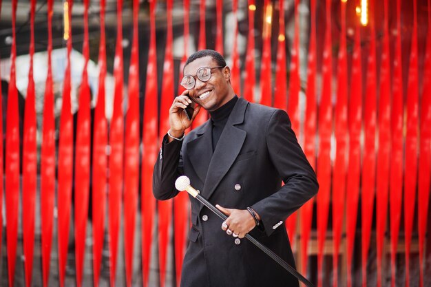 Stylish african american gentleman in elegant black jacket holding retro walking stick as cane flask or tippling cane Rich fashionable afro man against red wall speaking on mobile phone