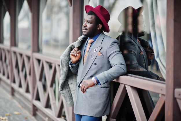 Stylish African American man model in gray coat jacket tie and red hat posed against wooden cafe