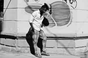 Free photo stylish african american man in white shirt and colored pants with hat and glasses posed outdoor against graffiti wall black fashionable model boy