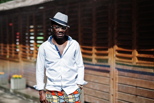 Stylish african american man in white shirt and colored pants with hat and glasses posed outdoor Black fashionable model boy