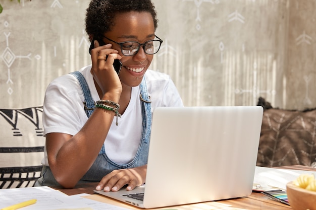 Stylish African American woman calls on smart phone looks in laptop computer, updates software