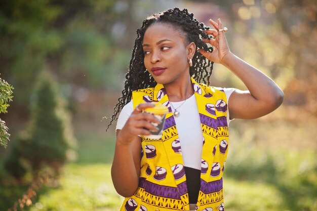 Stylish african american women in yellow jacket posed on street with hot drink in disposable paper cup