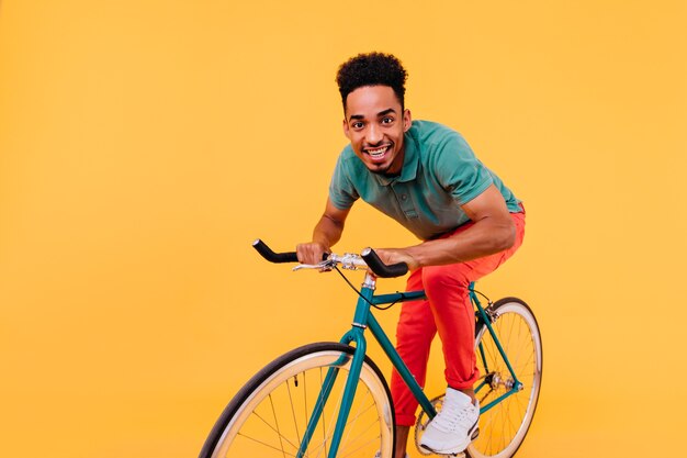 Stylish african guy in green t-shirt and white sneakers posing on bicycle. blissful black man riding on bike.