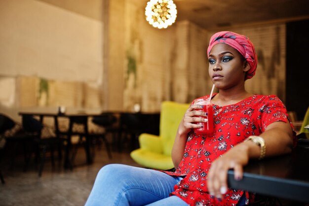 Stylish african woman in red shirt and hat posed indoor cafe and drinking strawberry lemonade