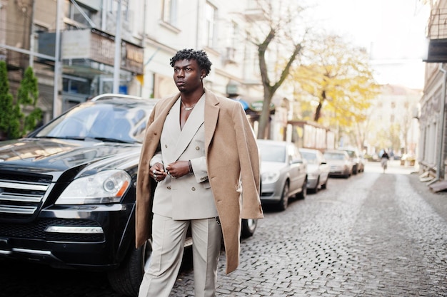 Stylish afro man in beige old school suit and coat walking against black business car Fashionable young African male in casual jacket on bare torso