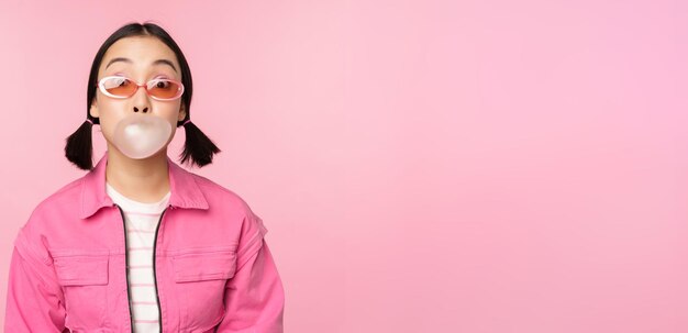 Stylish asian girl blowing bubblegum bubble chewing gum wearing sunglasses posing against pink background